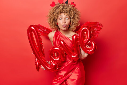 Studio portrait of young beautiful African american girl with curly short hair in red dress standing in centre holding sign love wanting to kiss you looking straight. St. Valentine's Day celebration