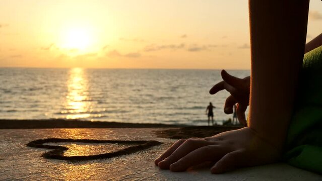 Close-up, a child's hand with a finger draws figures with sand on the concrete surface of the sidewalk on the beach embankment at sunset. Drawing hearts with sand using your fingers
