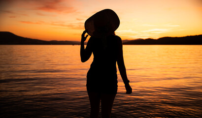 kobieta w kapeluszu na tle zachodzącego słońca nad morzem - obrazy, fototapety, plakaty