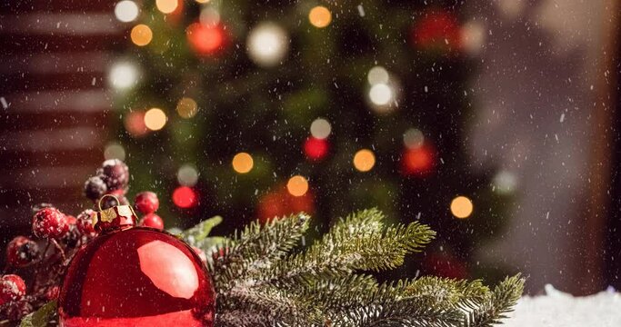 Animation of snow falling over bauble decoration against blurred view of christmas tree at home