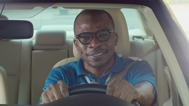 Medium portrait shot of positive African American driver sitting behind steering wheel and preparing to go somewhere by car, looking at camera