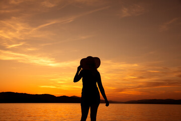 kobieta w kapeluszu na tle zachodzącego słońca nad morzem - obrazy, fototapety, plakaty