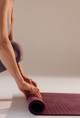 A young woman turns a mat before or after a yoga class in the studio. Close-up legs of a woman doing yoga while sitting on a light background - obrazy, fototapety, plakaty