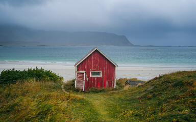 Red hut in the foreground next to a path leading to the Ramberg beach, Lofoten, Norway, during a cloudy day next to a mountains on the horizon.