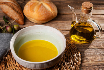 Bowl with olive oil next to an oil can and some bread rolls, with a rustic background.