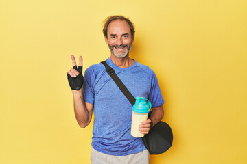 Gym-goer with protein shake in studio showing number two with fingers.