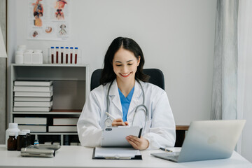 Confident young  Asian female doctor in white medical uniform sit at desk working on computer. Smiling use laptop write in medical journal