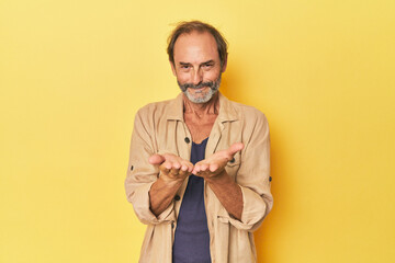 Caucasian middle-aged man in yellow studio holding something with palms, offering to camera.