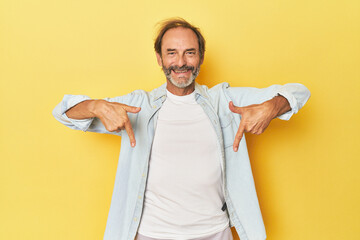 Caucasian middle-aged man in yellow studio points down with fingers, positive feeling.