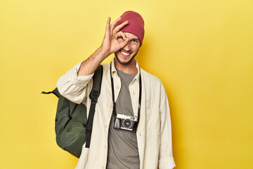 Man with vintage camera and backpack on yellow excited keeping ok gesture on eye.