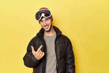 Man in winter coat with snow goggles on yellow pointing with finger at you as if inviting come closer.