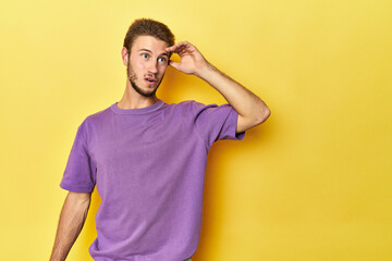 Young Caucasian man on a yellow studio background looking far away keeping hand on forehead.