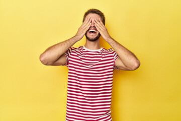 Young Caucasian man on a yellow studio background covers eyes with hands, smiles broadly waiting for a surprise.