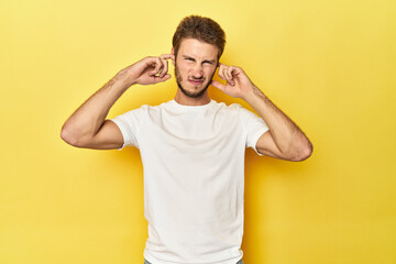 Young Caucasian man on a yellow studio background covering ears with fingers, stressed and desperate by a loudly ambient.