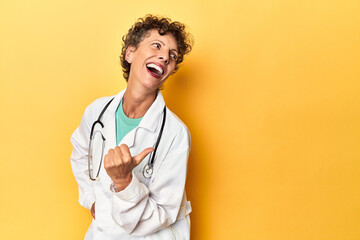 Caucasian mid-age female doctor on yellow studio points with thumb finger away, laughing and...