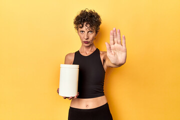 MIddle aged athlete woman holding protein supplement on yellow standing with outstretched hand showing stop sign, preventing you. - obrazy, fototapety, plakaty