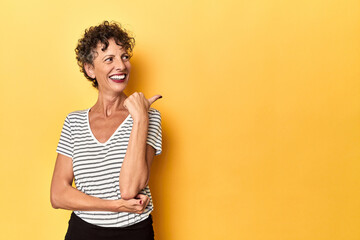 Mid-aged caucasian woman on vibrant yellow points with thumb finger away, laughing and carefree.