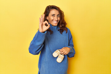 Pregnant woman holding baby shoes on yellow cheerful and confident showing ok gesture.
