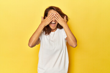 Middle-aged woman on a yellow backdrop covers eyes with hands, smiles broadly waiting for a surprise.