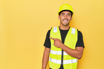 Latino worker in yellow vest and helmet smiling and pointing aside, showing something at blank space.