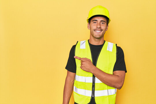 Latino worker in yellow vest and helmet smiling and pointing aside, showing something at blank space.