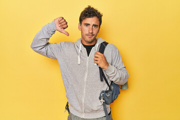 Young Latino with mountain backpack on yellow showing a dislike gesture, thumbs down. Disagreement...