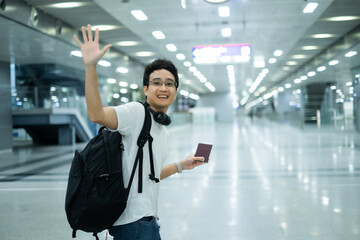Young Asian millenael man standing with smile in air port or rail station wave his hand to someone...