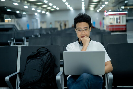 Young Asian millenael man sitting and working with laptop in air port or rail station while waiting for train or airplain with black backpack and headphone.