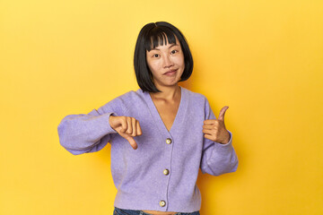 Young Chinese lady, yellow studio background showing thumbs up and thumbs down, difficult choose...