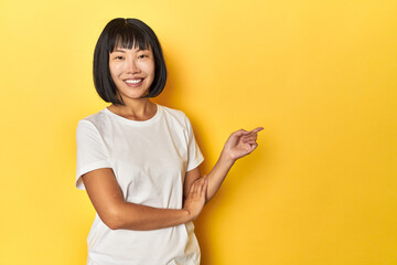 Young Chinese lady, yellow studio background smiling cheerfully pointing with forefinger away.