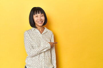 Young Chinese lady, yellow studio background smiling and pointing aside, showing something at blank...