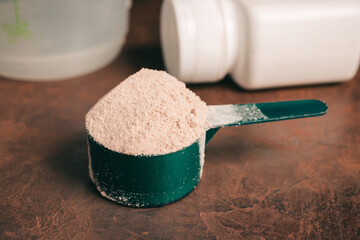 Chocolate whey protein powder in measuring spoon, old rusty dumbbell, shaker, plastic bottle on...
