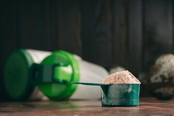 Chocolate whey protein powder in measuring spoon, old rusty dumbbell, shaker, plastic bottle on...