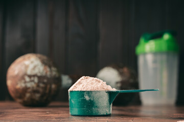 Chocolate whey protein powder in measuring spoon, old rusty dumbbell and shaker on dark background....