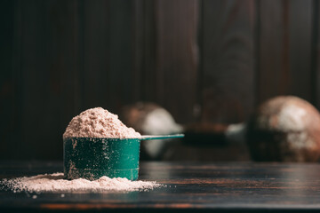 Chocolate whey protein powder in measuring spoon, old rusty dumbbell on dark background. healthy...
