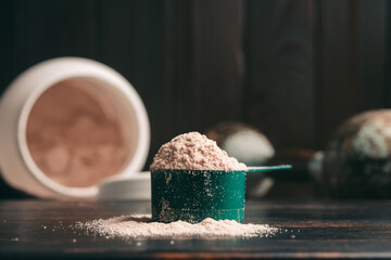 Chocolate whey protein powder in measuring spoon, old rusty dumbbell, plastic jar on dark...