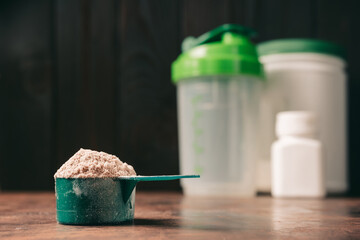 Chocolate whey protein powder in measuring spoon, old rusty dumbbell, shaker, plastic bottle on dark background. healthy eating, bodybuilding food supplements