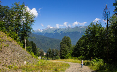 A beautiful panoramic landscape - a walking path among mountains and green trees on a sunny summer day and a blue sky with white clouds. Concept hiking and trekking