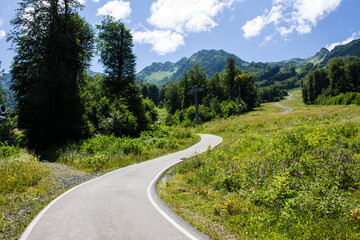 A beautiful panoramic landscape - a walking path among mountains and green trees on a sunny summer day and a blue sky with white clouds. Concept hiking and trekking