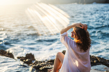 Young cheerful relaxed woman in a white shirt enjoying a wonderful moment of life while sitting on the seashore at sunset - obrazy, fototapety, plakaty