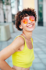 Fototapeta na wymiar Young beautiful smiling hipster woman in trendy summer clothes. Carefree woman with curls hairstyle, posing in the street at sunny day. Positive model outdoors. Cheerful and happy. In sunglasses