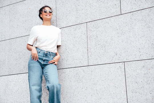 Young beautiful smiling hipster woman in trendy summer white t-shirt and jeans clothes. Carefree woman, posing in the street at sunny day. Positive model outdoors near wall. in sunglasses