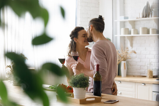 Happy peaceful young couple enjoying romantic dating in home kitchen, drinking wine, cooking dinner, hugging with love, care, tenderness, kissing at table with bottle, healthy food ingredients
