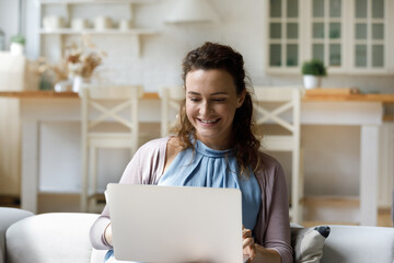 Happy young freelance worker woman working at laptop computer from home couch, typing, using online virtual app on digital device for communication, shopping on Internet, chatting, studying
