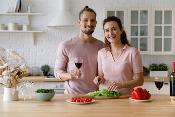 Cheerful happy young couple of bloggers cooking dinner in kitchen head shot portrait. Millennial husband and wife drinking wine over preparing salad, cutting fresh vegetables, smiling at camera