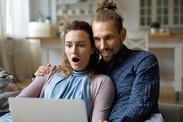 Shocked happy young married couple getting email message with surprising good news, looking at...