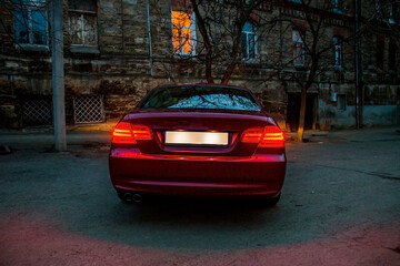 The trunk and glowing headlights of a red car against the background of a house