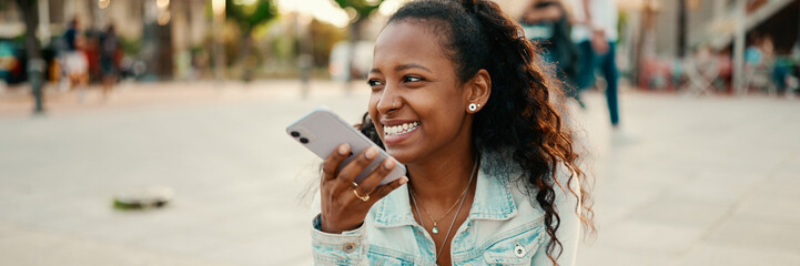 Closeup portrait of young woman with long curly hair using a mobile phone in an urban city background. Close-up of  girl sending a voice message on a smartphone
