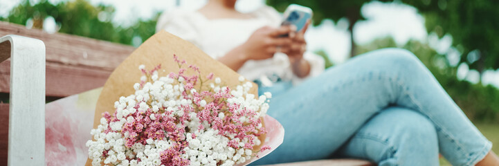 Young woman sits in the park on a bench with a bouquet of flowers with a smartphone in her hands....