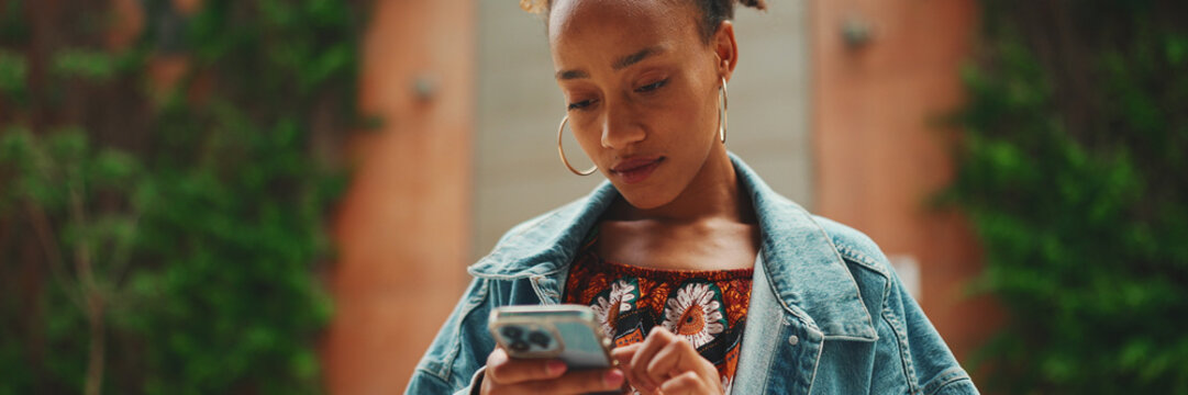 Close-up African girl wearing denim jacket, goes on the street of modern city and holding smartphone looking at phone screen look for address with electronic map in city